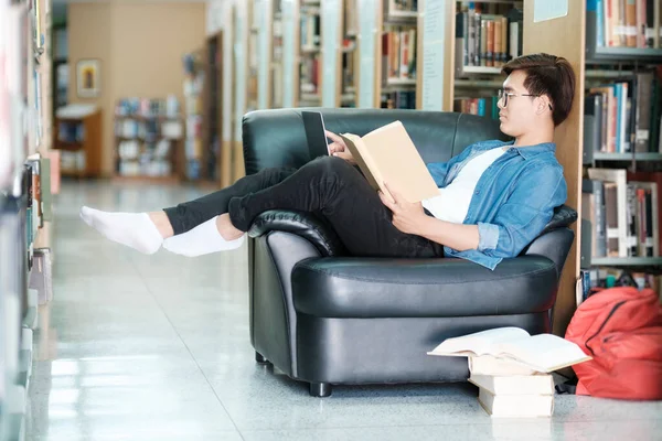 Young male college student wearing eyeglasses and casual clothings sitting on couch reading book, studying and doing research for school project at a library. E-Learning and Educational concept.