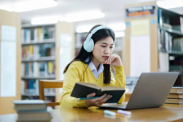 若い女子大生 ヘッドフォンを装着した高校生 机の読書本に座っているカジュアルな服で 図書館で学校プロジェクトのためのラップトップを使用して研究を勉強し 研究を行っています Learning Educationの概念 — ストック写真