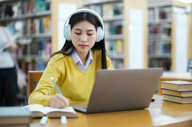 Genç bayan kolej, kulaklık takan lise öğrencisi ve günlük kıyafetleri olan bir kütüphanede dizüstü bilgisayarla ders çalışıp araştırma yapan bir öğrenci. E-Öğrenim Eğitim Kütüphanesi konsepti.