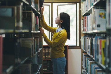 Asyalı genç bayan öğrenci ayakta duruyor, kütüphanede okumak için kitap arıyor ve seçiyor, akademik araştırma, ya da okul çalışması ya da proje için. Öğrenim, Eğitim, Kütüphane konsepti.