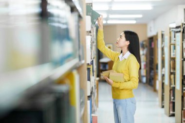 Asyalı genç bayan öğrenci ayakta duruyor, kütüphanede okumak için kitap arıyor ve seçiyor, akademik araştırma, ya da okul çalışması ya da proje için. Öğrenim, Eğitim, Kütüphane konsepti.
