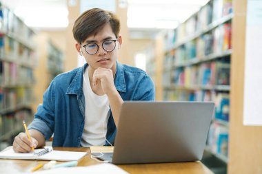 Gözlüklü genç bir üniversite öğrencisi masa başında oturmuş ders çalışıyor, kitap okuyor, çok düşünüyor ve araştırma için kütüphanede kulaklık takarak bilgisayar kullanarak notlar yazıyor.