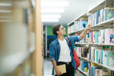 Sırt çantalı, ayakta duran, kütüphanede okumak, akademik araştırma ya da okul ödevi için kitap seçen genç bir erkek öğrenci. Öğrenme, Eğitim konsepti.