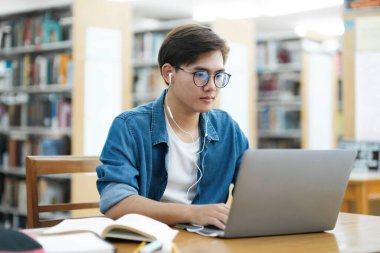 Kolej öğrencisi gözlük ve kulaklık takıyor ve günlük giysiler içinde masa başında oturmuş kütüphanede dizüstü bilgisayarla araştırma yapıyor ve kitap okuyor. Çevrimiçi eğitim, E-Öğrenme