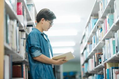 Sırt çantalı, ayakta duran, kitap okuyan ya da okumak için kütüphanede kitap seçen, akademik araştırma ya da okul ödevi yapan genç bir erkek öğrenci. Öğrenme, Eğitim konsepti.