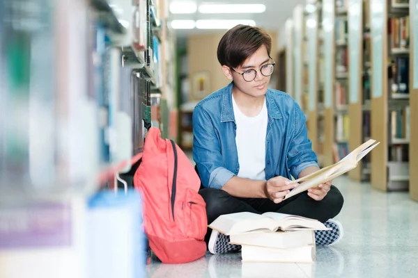 年轻的男大学生戴着眼镜 身穿休闲装 坐在地板上看书 在一所图书馆为学校的项目做研究 学习和教育概念 — 图库照片