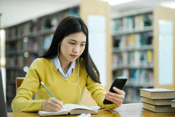 年轻的女大学生 戴着耳机 穿着休闲装 坐在课桌前学习 在图书馆用手机写作 以进行研究或实施学校项目 电子学习 — 图库照片