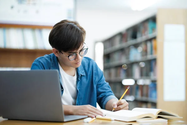 年轻的男大学生戴着眼镜 穿着休闲装 在图书馆的书桌上用笔记本电脑学习 阅读和记笔记 为学校的项目或考试做准备 — 图库照片