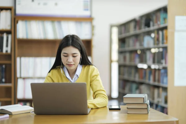 若い女子大生 ヘッドフォンを装着した高校生 机の読書本に座っているカジュアルな服を着て 図書館で学校プロジェクトのためのラップトップを使用して研究を勉強し 研究を行っています Learning 教育図書館構想 — ストック写真