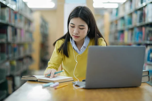 年轻的女大学生 戴着耳机 穿着休闲装坐在写字台前 坐在笔记本上 在图书馆用笔记本电脑学习和做研究 电子学习教育图书馆概念 — 图库照片
