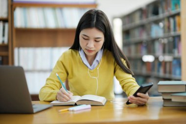 Genç bayan kolej, kulaklık takan lise öğrencisi yerde oturmuş kitap okuyor ve bir kütüphanedeki okul projesi için dizüstü bilgisayar ve telefon kullanarak araştırma yapıyor. E-Öğrenim Eğitim Kütüphanesi konsepti.