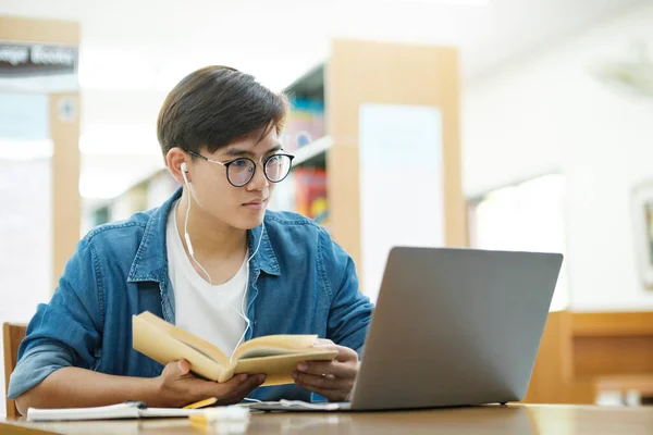 眼鏡をかけ 机に座って勉強したり 研究や学校のプロジェクトのために図書館でヘッドフォンをしてノートパソコンを使って本を読んだりする若い男性大学生 Learningの概念 — ストック写真