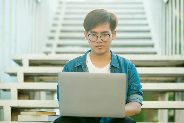 Young male college student with eyeglasses and in casual clothings smiling and sitting outdoor on stairs to study using laptop looking for information for school project and research. Education and E-Learning concept.