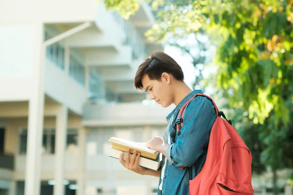 年轻聪明的男大学生穿着休闲装 背着背包站在室外 背景是校园大楼 手里拿着和阅读着准备在课堂上学习的书籍 教育概念 — 图库照片