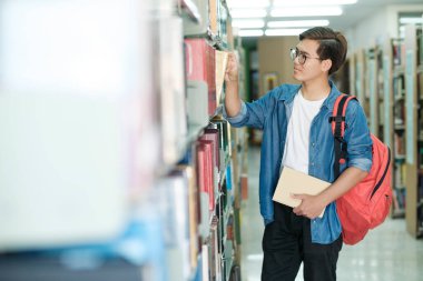 Sırt çantalı, ayakta duran, kütüphanede okumak, akademik araştırma ya da okul ödevi için kitap seçen genç bir erkek öğrenci. Öğrenme, Eğitim konsepti.