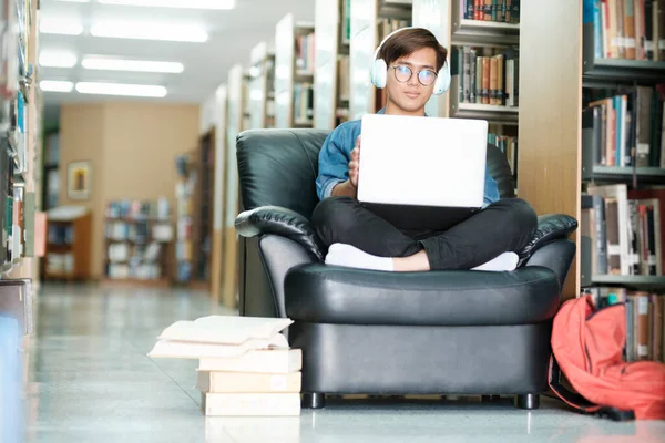 眼鏡をかけ ソファに座って勉強したり 研究や学校のプロジェクトのために図書館でヘッドフォンを着用したラップトップを使用している若い男性大学生 Learning Educationの概念 — ストック写真
