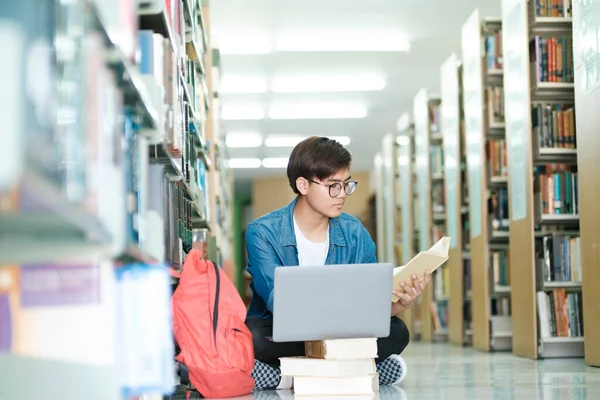 年轻的男大学生戴着眼镜和休闲装坐在地板上看书 在图书馆用笔记本电脑学习和做研究 学习和教育概念 — 图库照片