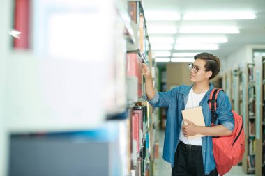 Sırt çantalı, ayakta duran, kütüphanede okumak, akademik araştırma ya da okul ödevi için kitap seçen genç bir erkek öğrenci. Öğrenme, Eğitim konsepti.