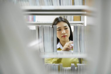 Asyalı genç bayan öğrenci ayakta duruyor, kütüphanede okumak için kitap arıyor ve seçiyor, akademik araştırma, ya da okul çalışması ya da proje için. Öğrenim, Eğitim, Kütüphane konsepti.