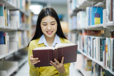 Gündelik giysiler içinde genç bir üniversite öğrencisi yerde oturmuş kitap okuyor ve kütüphanede okul projesi için keyif alarak araştırma yapıyor. Öğrenim, Eğitim, Kütüphane konsepti.