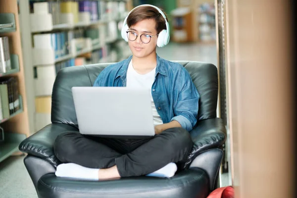 年轻的男大学生戴着眼镜 穿着休闲装 坐在沙发上 并在图书馆使用笔记本电脑 带着耳机进行研究或实施学校项目 电子学习和教育概念 — 图库照片