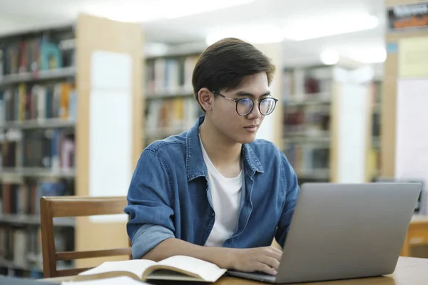Young male college student wearing eyeglasses and in casual cloths sitting at desk to study and read books using laptop at library to searcg for informationfor research or school project. E-learning and Education concpet.