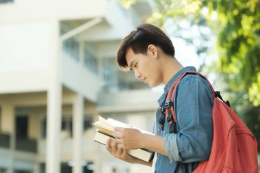 Sırt çantası ve günlük giysiler içinde, arka planda kampüs binası olan genç ve zeki erkek kolaj öğrencisi, elinde kitap tutuyor ve derste okumaya hazır. Eğitim kavramı.