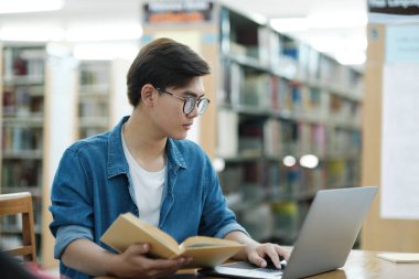 Genç üniversite öğrencisi gözlük takıyor ve gündelik giysiler içinde kütüphanede araştırma ya da okul projesi hakkında bilgi aramak için dizüstü bilgisayarla ders çalışmak ve okumak için masa başında oturuyor. E-öğrenim ve Eğitim kavramı.