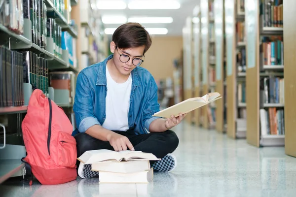 Young male college student wearing eyeglasses and casual clothings sitting on the floor reading book, studying and doing research for school project at a library. Learning and Educational concept.