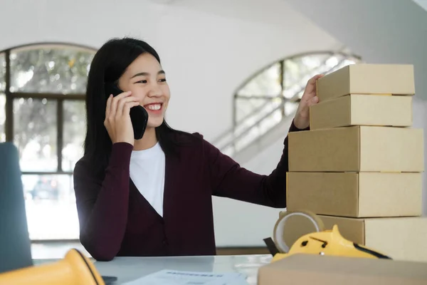 Young asian female online business owner or entrepreneur with smiley face sitting at desk with boxes, laptop, scanner, and taper talking to customer for order using smartphone. Online business, e-commerce concept.