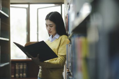 Ayakta duran, okumak ya da okumak için kütüphanede kitap seçen akademik araştırma, okul ödevi, proje ya da sınav yapan genç bir üniversite öğrencisi. Öğrenim, Eğitim, Kütüphane konsepti.