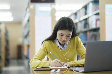 Genç bayan kolej, kulaklık takan lise öğrencisi ve günlük kıyafetleri olan bir kütüphanede dizüstü bilgisayarla ders çalışıp araştırma yapan bir öğrenci. E-Öğrenim Eğitim Kütüphanesi konsepti.