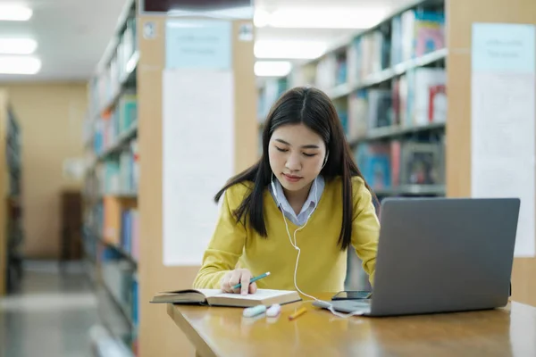 年轻的女大学生 戴着耳机 穿着休闲装坐在写字台前 坐在笔记本上 在图书馆用笔记本电脑学习和做研究 电子学习教育图书馆概念 — 图库照片