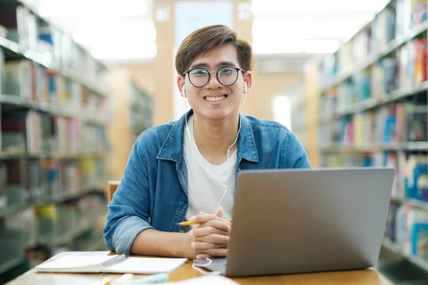 眼鏡をかけ机の上に座ってカメラを見ながら 研究や学校のプロジェクトのためにノートパソコンを使って本を勉強したり読んだりする若い男性大学生 学びの概念 — ストック写真
