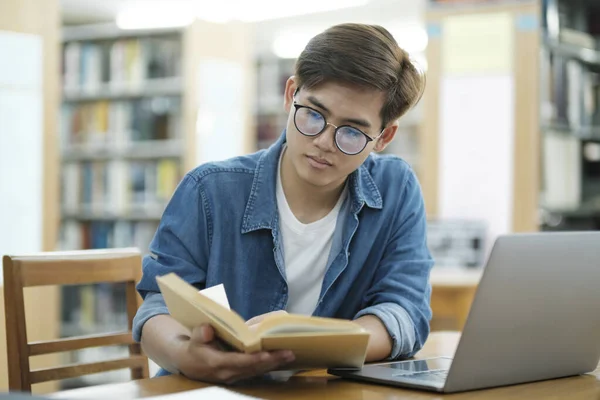 年轻的男大学生戴着眼镜 穿着休闲装 在图书馆用笔记本电脑学习和看书 以进行研究或实施学校项目 电子学习和教育概念 — 图库照片