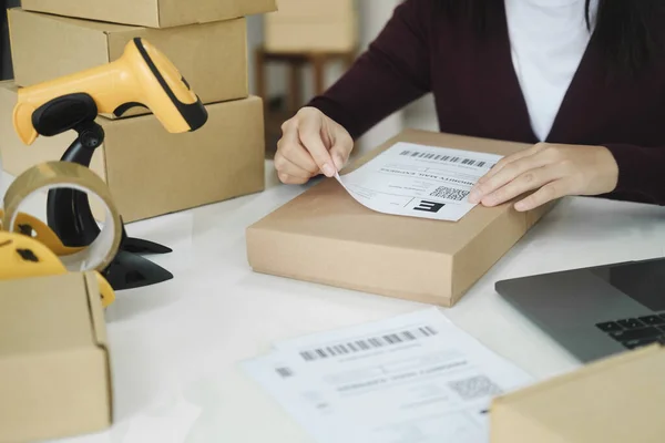 Close up of young female online business owner sitting at desk with laptop and boxes and putting shipping label on cardboard boxes or parcel of customer\'s order preparing for shipment and delivery. Online business, e-commerce, logistics concept.