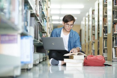 Genç erkek kolej, gözlük takan lise öğrencisi ve günlük giysiler giymiş yerde oturmuş kitap okuyor ve kütüphanedeki okul projesi için dizüstü bilgisayarla araştırma yapıyor. E-Öğrenme ve Eğitim Konsepti.