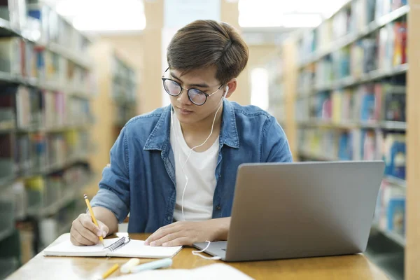 年轻的男大学生戴着眼镜 身穿休闲装 坐在课桌前学习 用笔记本电脑写笔记 带着耳机参加研究或学校项目 学习概念 — 图库照片