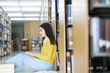 Genç bayan kolej, günlük giysiler içinde lise öğrencisi yerde oturmuş kitap okuyor ve kütüphanedeki okul projesi için dizüstü bilgisayarla araştırma yapıyor. E-Öğrenme ve Eğitim