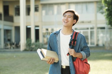 Sırt çantası ve günlük kıyafetlerle gezen zeki, erkek kolaj öğrencisi arka tarafta kampüs binasıyla ayakta duruyor ve eğitilmek için yukarı bakarken kitapları tutuyor. Eğitim