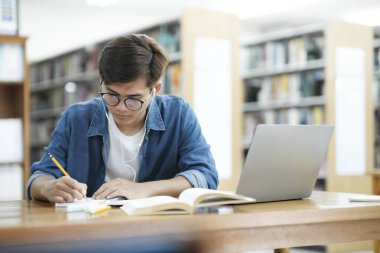 Gözlüklü ve günlük kıyafetlerle genç bir üniversite öğrencisi çalışma masasında oturuyor, kitap okuyor ve araştırma ya da okul için kütüphanede kulaklık takarak dizüstü bilgisayar kullanarak notlar yazıyor.