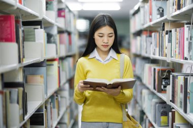 Ayakta duran, okumak ya da okumak için kütüphanede kitap seçen akademik araştırma, okul ödevi, proje ya da sınav yapan genç bir üniversite öğrencisi. Öğrenme, Eğitim