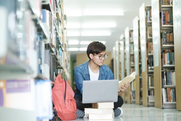 年轻的男大学生戴着眼镜和休闲装坐在地板上看书 在图书馆用笔记本电脑学习和做研究 学习和教育 — 图库照片