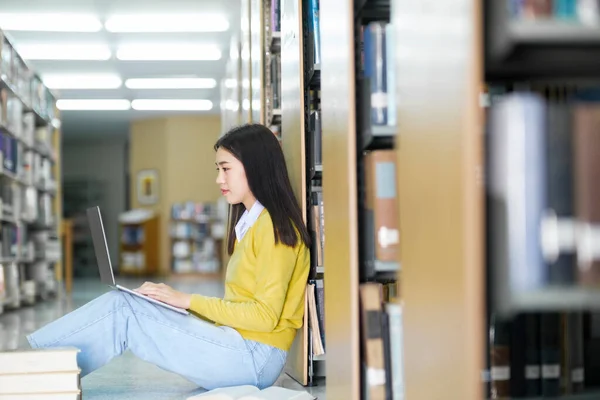 年轻的女大学生 穿着休闲装坐在地板上看书 在图书馆用笔记本电脑学习和做研究 电子学习和教育 — 图库照片