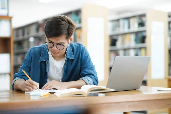 眼鏡をかけ 机に座って勉強したり 本を読んだり 研究や学校のために図書館でヘッドフォンをしてノートを書き留めたりする若い男性大学生 — ストック写真
