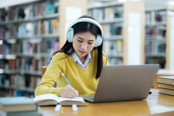 年轻的女大学生 戴着耳机 穿着休闲装坐在写字台前 坐在笔记本上 在图书馆用笔记本电脑学习和做研究 — 图库照片