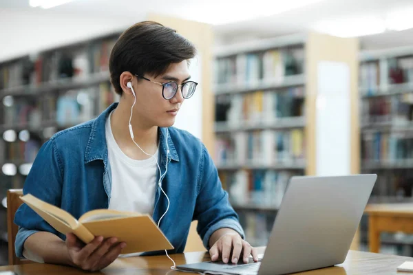 年轻的男大学生 戴着眼镜和耳机 身穿休闲装 坐在课桌前学习 并在图书馆用笔记本电脑进行研究或实施学校项目 — 图库照片