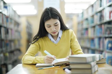 Gündelik giysiler içinde genç bir üniversite öğrencisi çalışma masasında oturuyor, kitap okuyor ve kütüphanede araştırma ya da okul projesi için sadece altını çiziyor. E-Öğrenme Eğitim Kütüphanesi