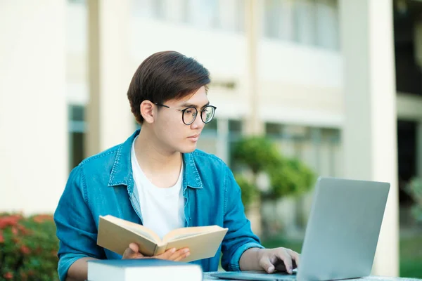 年轻的男大学生 戴着眼镜 身穿休闲装 坐在室外学习 读书和看书 他们使用笔记本电脑搜寻学校项目和研究的信息 教育和培训 — 图库照片