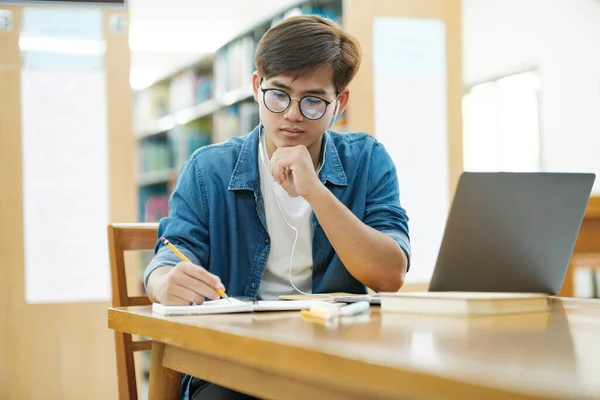 Young male college student wearing eyeglasses and in casual cloths sitting at desk studying, reading book, thinking hard, and writing down notes using laptop wearing headphones at library for research